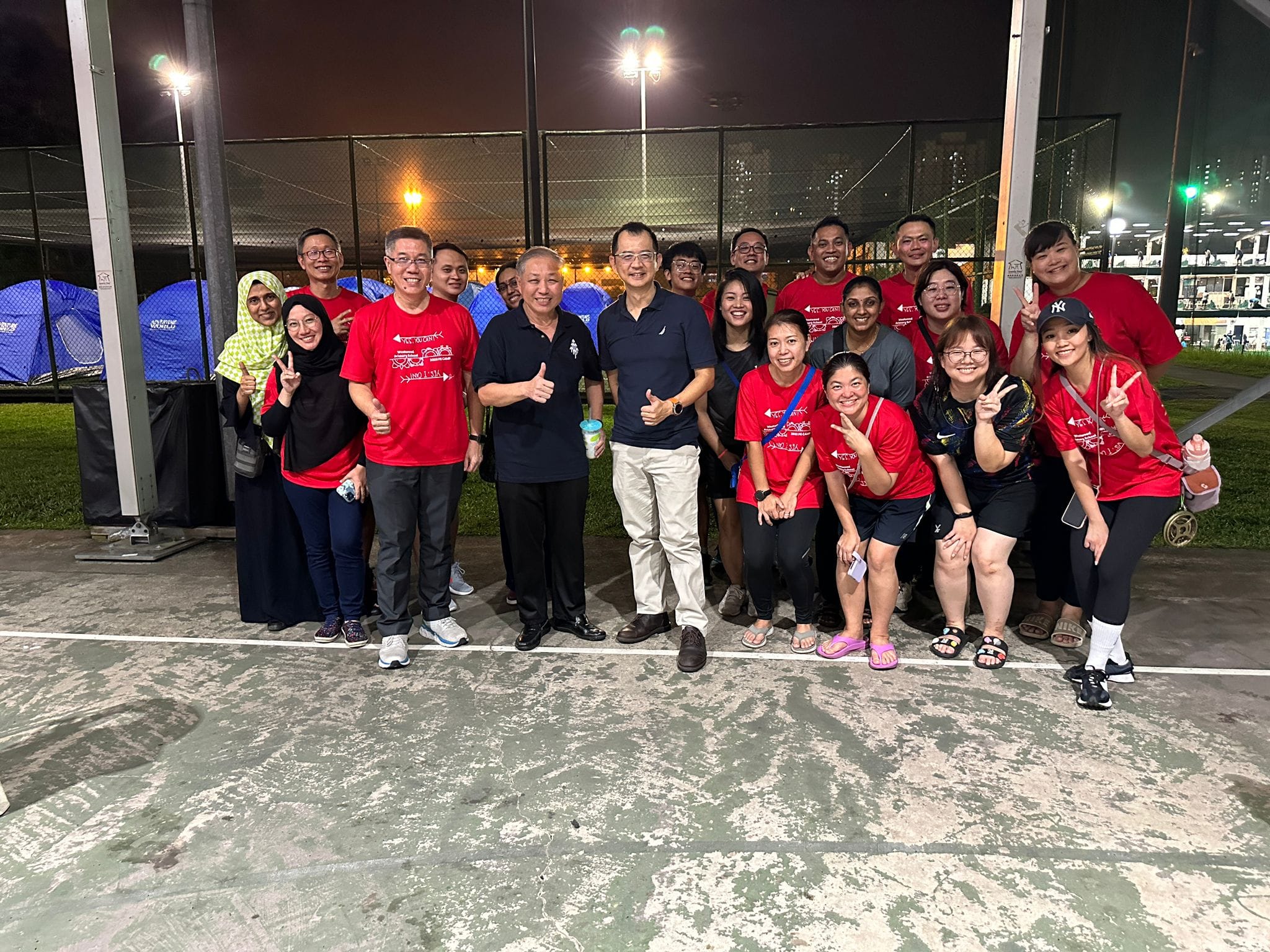 A group of about 20 people posing at a lit outdoor sports court at night, most wearing red shirts.
